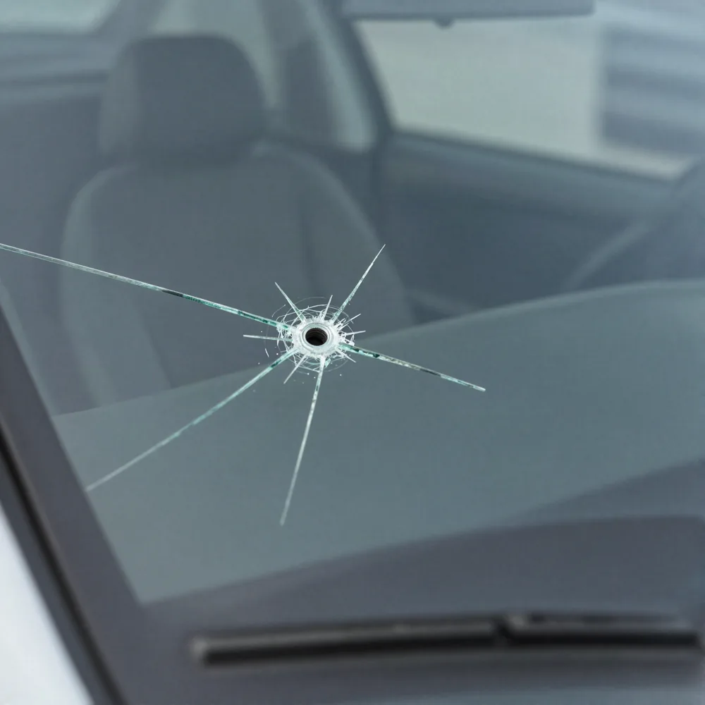 A bullseye stone chip with crack legs spreading through a car windshield, dashboard visible behind the glass